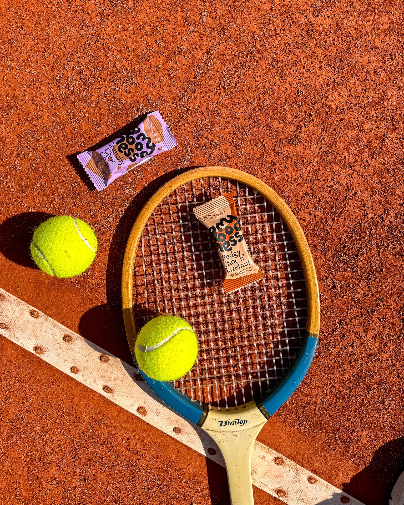 My Goodness Crunchy Choc Chip and Fudgy Choc n' Hazelnut bars on a vintage tennis racket and clay court — the perfect on-the-go snack